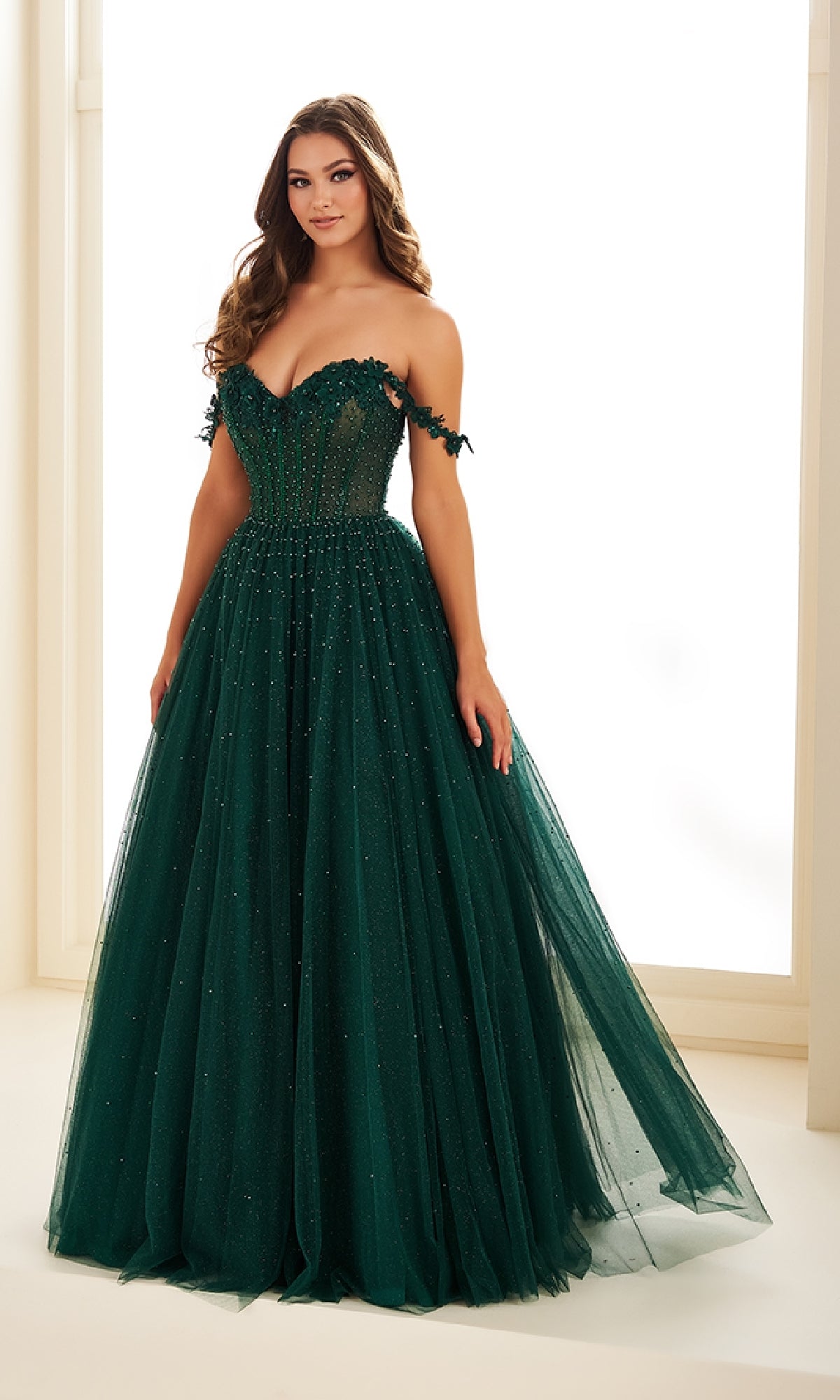 A woman stands indoors wearing the Ellie Wilde EW36071, a long prom dress with an off-the-shoulder fitted bodice and flowing tulle skirt in dark green.